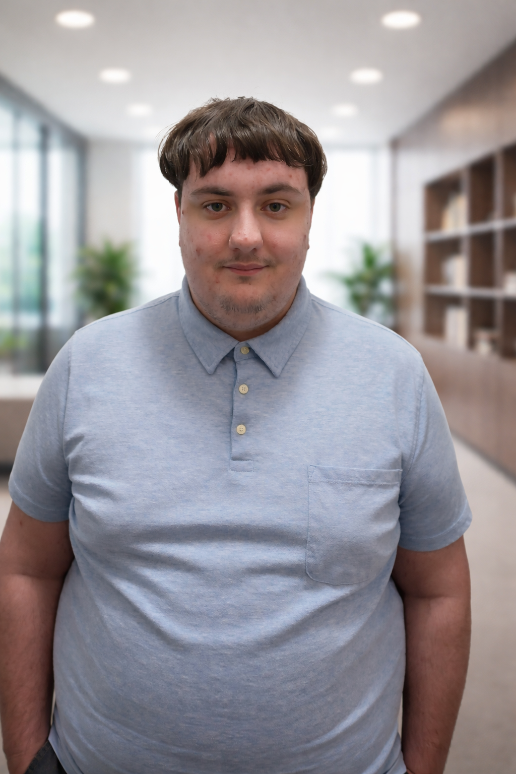 Portrait of a person wearing a light blue polo shirt, standing in a modern office environment with a softly blurred background.